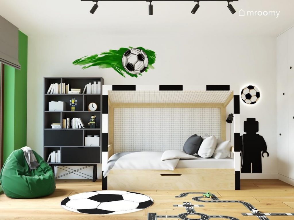Drewniane łóżko w formie bramki do piłki nożnej a obok szary regał zielona pufa oraz tablica kredowa w kształcie ludzika Lego a na podłodze i ścianach piłki