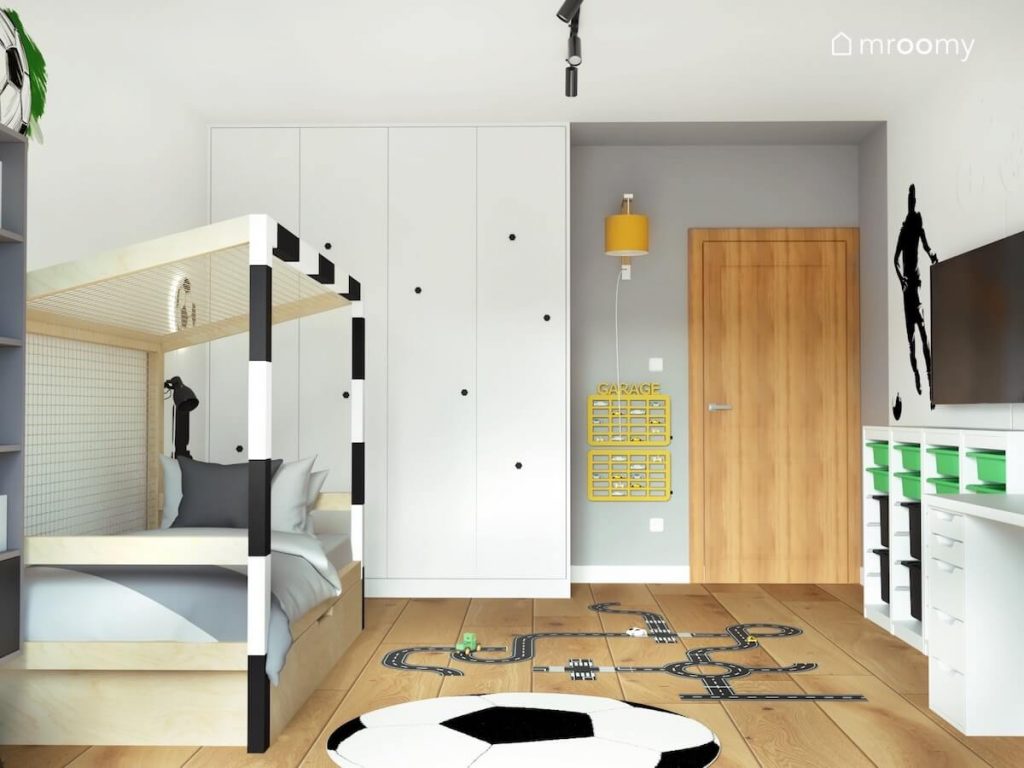 Biało szary pokój chłopca w wieku szkolnym a w nim drewniane łóżko w formie bramki duża biała szafa dywan piłka naklejka podłogowa w kształcie jezdni oraz żółte półki na autka