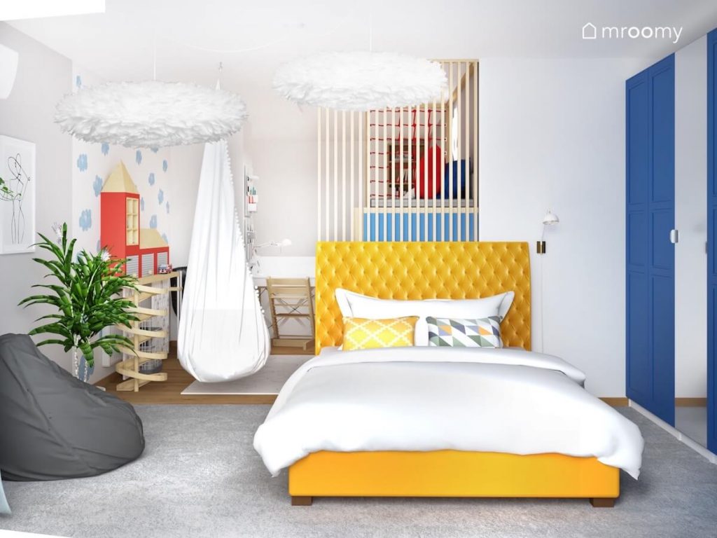 Duże żółte łóżko w pokoju kilkulatka a obok szara pufa a na suficie lampy z piór