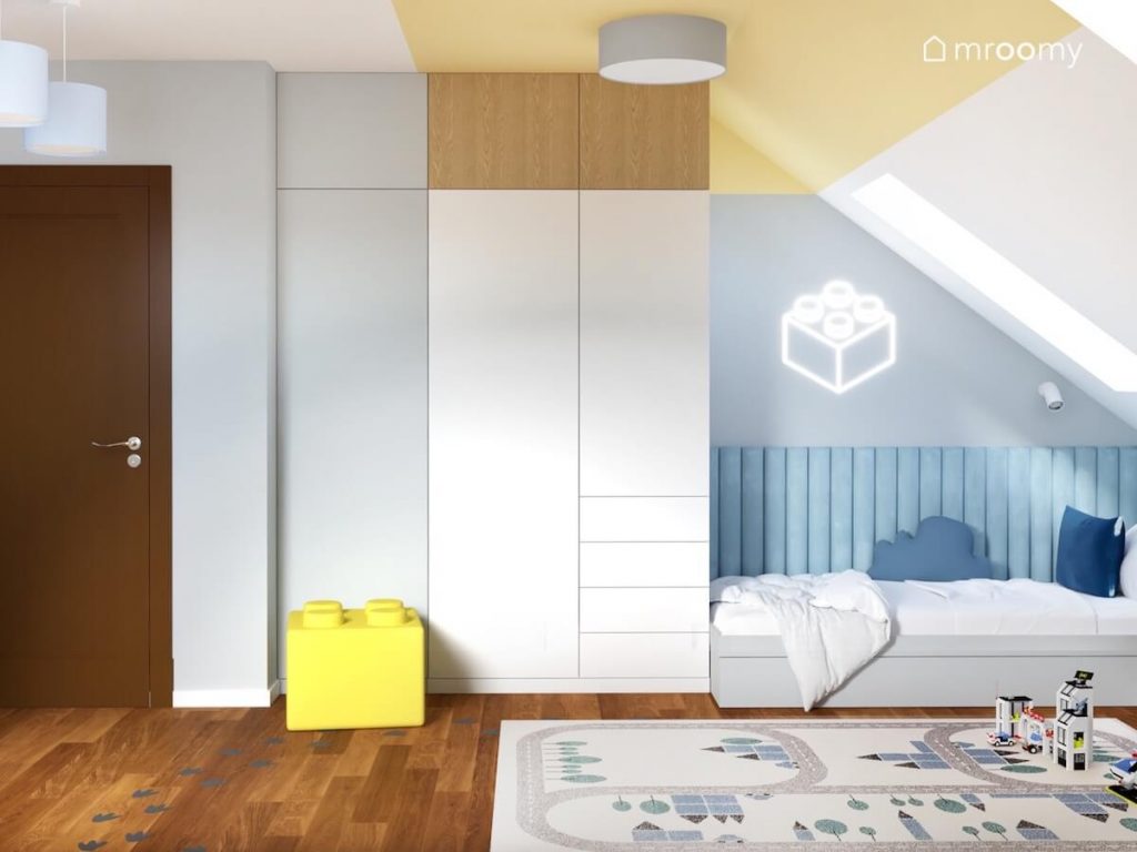 Jasny pokój dwóch chłopców a w nim biała szafa z drewnianym elementem szare łóżko uzupełnione niebieskimi panelami ściennymi oraz ledon i pufka w kształcie klocka