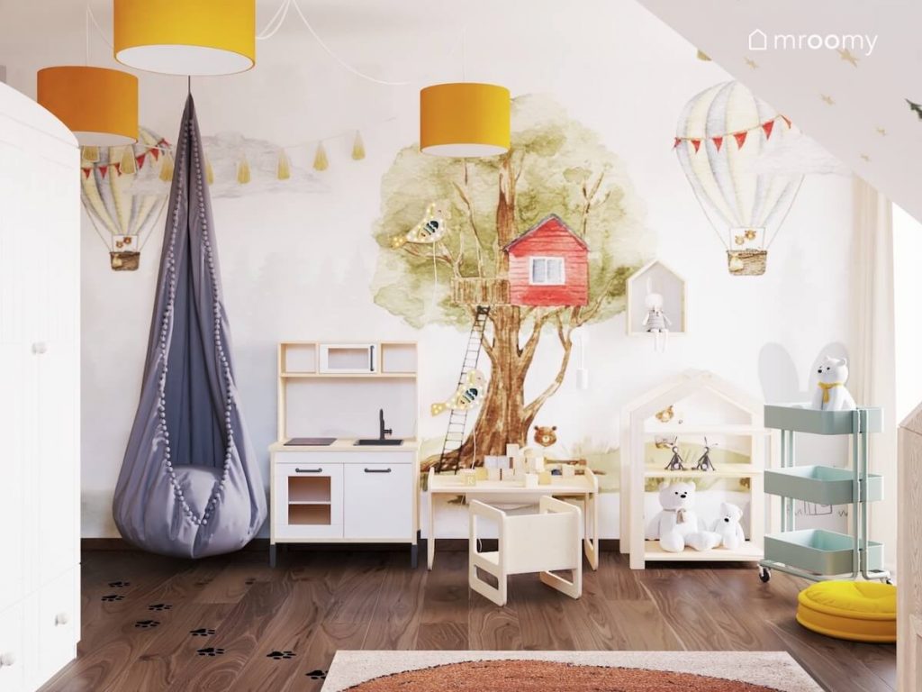 Ściana w pokoju dziewczynki pokryta tapetą z domkiem na drzewie i balonami a także kuchenka dla dzieci oraz drewniany stoliczek do zabawy a na suficie fotel kokon i żółte lampy