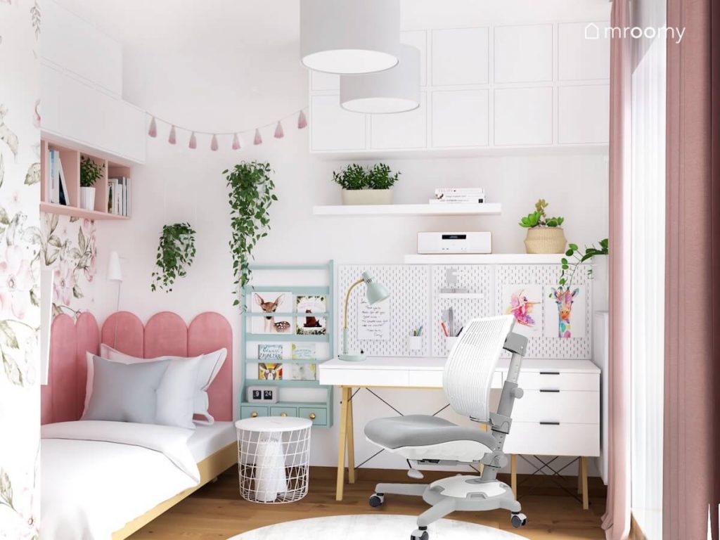 Biały pokój dla dziewczynki a w nim drewniane łóżko uzupełnione różowymi panelami biurko z kontenerkiem i szarym krzesłem błękitna biblioteczka oraz wiele kwietników