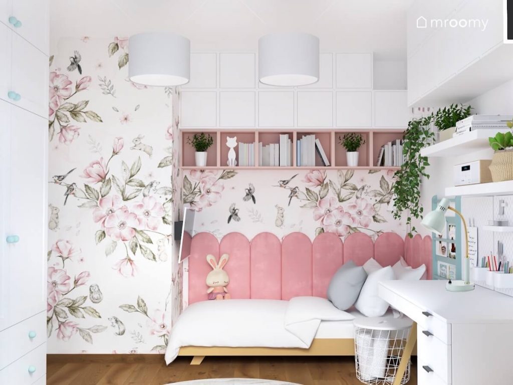 Ściana w pokoju pięciolatki ozdobiona tapetą w kwiaty a na niej biało różowe szafki oraz łóżko uzupełnione różowymi panelami w kształcie zaokrąglonych sztachet