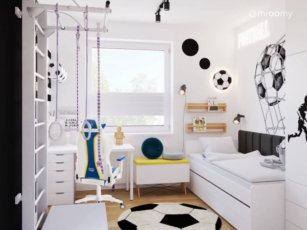 Jasny pokój dla fana piłki nożnej a w nim białe meble kolorowy fotel gamingowy oraz motywy piłkarskie w postaci lampki ściennej naklejek dywanu i ledonu