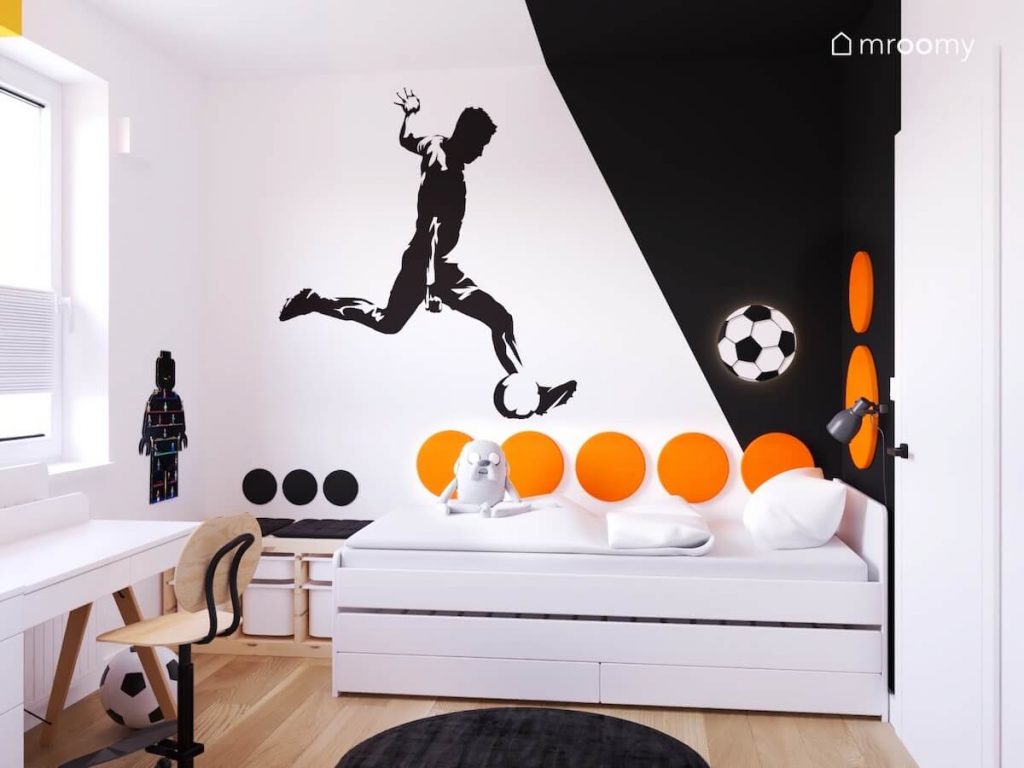 Biało czarna ściana w pokoju chłopca a na niej naklejka z piłkarzem lampka nocna w kształcie piłki oraz okrągłe pomarańczowe panele ścienne
