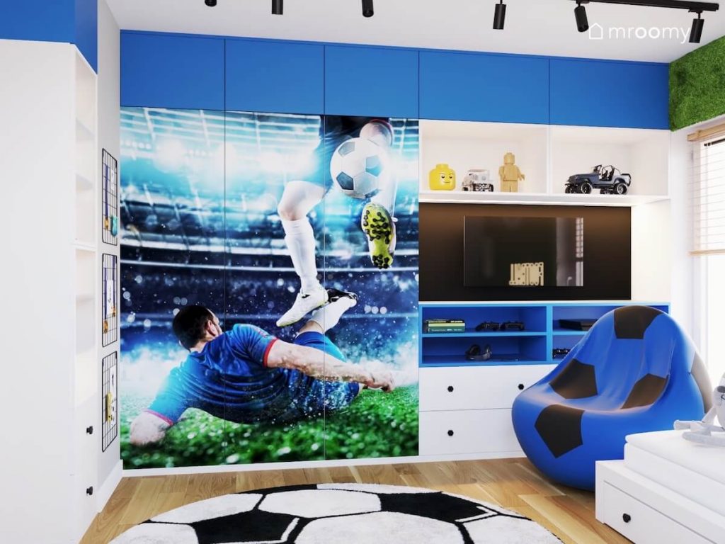 Biało niebieska zabudowa meblowa a między nią telewizor a na trzydrzwiowej ścianie piłkarska fototapeta w pokoju chłopca