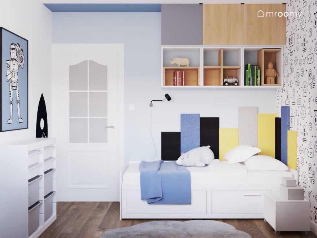 Białe łóżko w pokoju chłopca uzupełnione panelami ściennymi w różnych kolorach a nad nim szafki w różnych kolorach a obok biały regał z pojemnikami i plakat z robotem