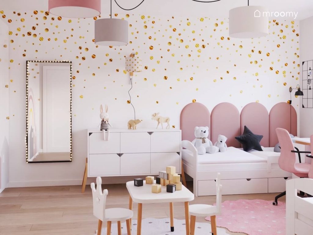 Biała ściana w złote kropki w pokoju dwóch dziewczynek a na niej prostokątne lustro biała komoda na drewnianych nogach oraz białe łóżko z różowymi panelami