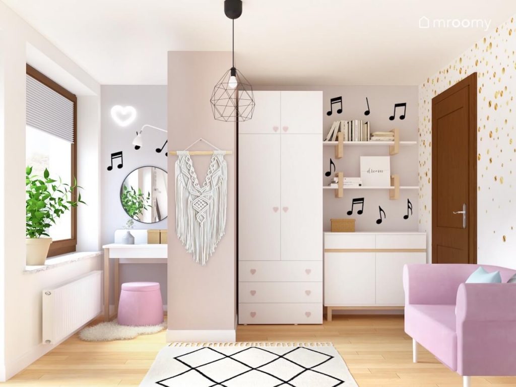 Toaletka z okrągłym lustrem różową pufą i miękkim dywanikiem oraz biała szafa z gałkami w kształcie serduszek a na ścianach półki na książki makrama i naklejki nutki w pokoju dziewczynki