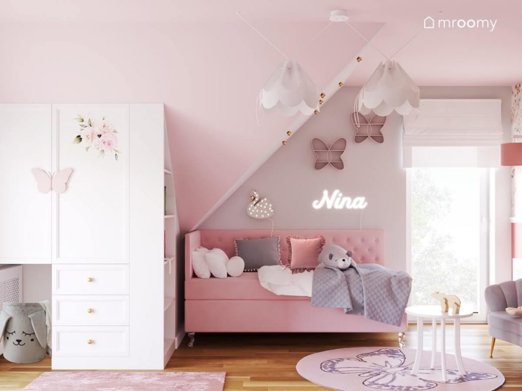 Różowo szary poddaszowy pokój dla dziewczynki a w nim biała szafa ze złotymi gałkami różowe tapicerowane łóżko oraz półeczki w kształcie motyli