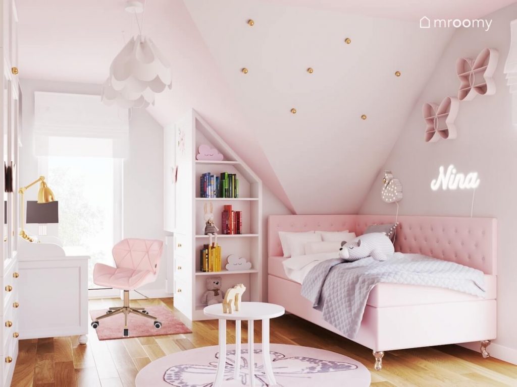 Jasny poddaszowy pokój dla dziewczynki a w nim geometryczny regał różowe tapicerowane łóżko dywan z motylem oraz skos ozdobiony złotymi gałkami