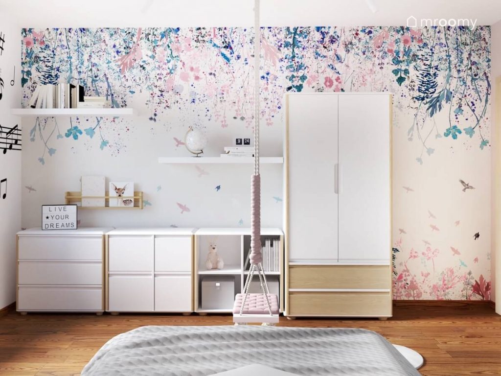 Ściana w pokoju dziewczynki pokryta tapetą w kwiaty a na niej biało drewniana szafa półki biblioteczka i niskie meble a u sufitu huśtawka w frędzlami