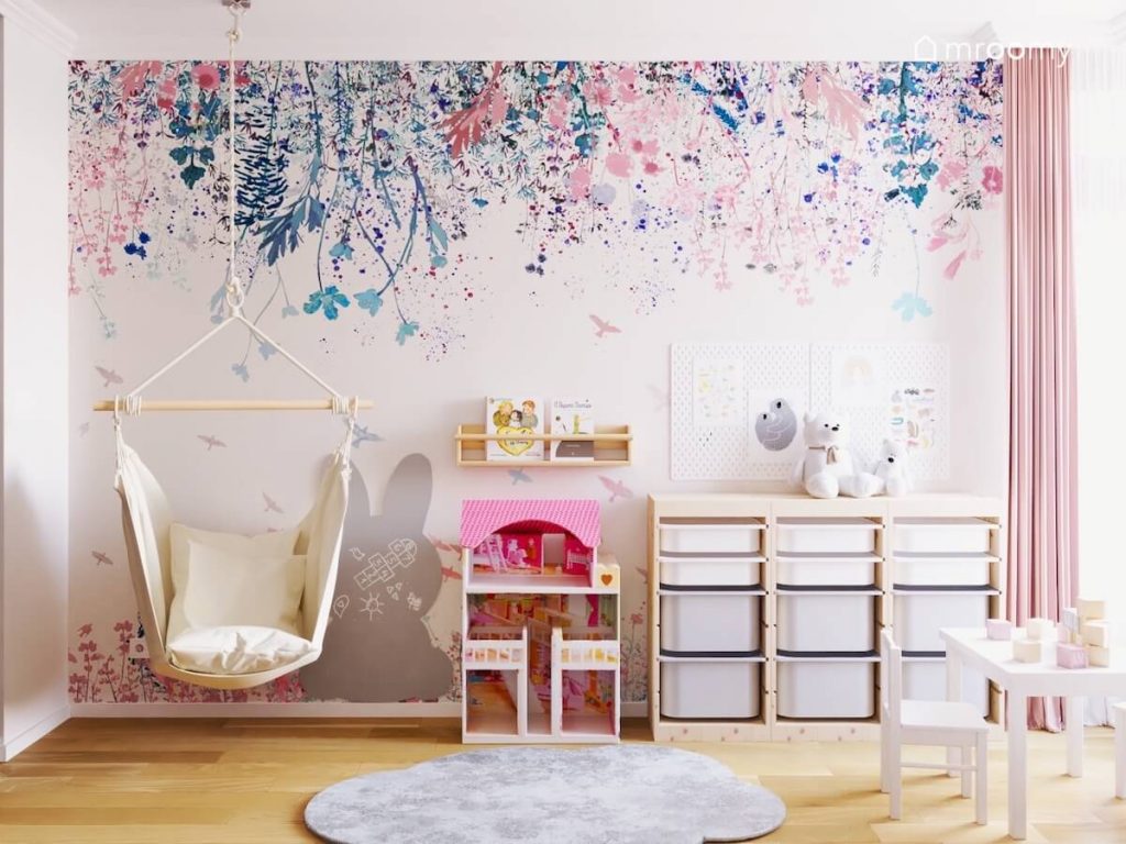 Kremowy fotel wiszący tablica kredowa w kształcie królika domek dla lalek oraz drewniany regał z pojemnikami a na ścianie tapeta w kwiaty w pokoju małej dziewczynki