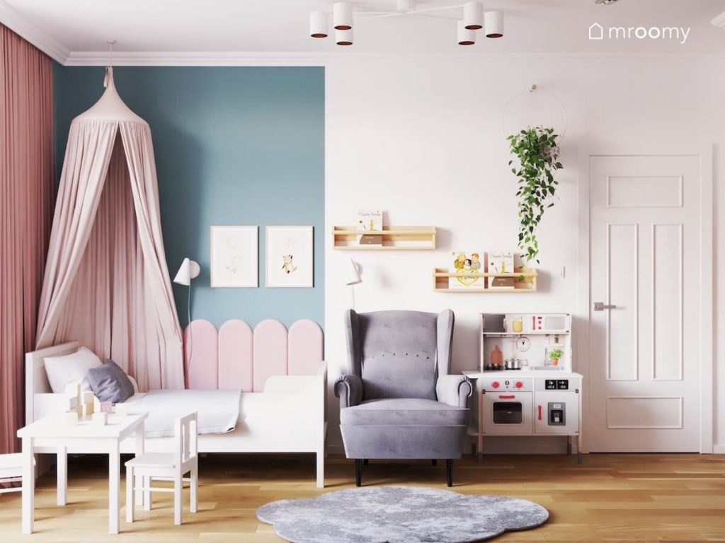 Niebiesko biała ściana w pokoju trzylatki a na niej drewniane półeczki kwietnik oraz ozdobne plakaty a poniżej łóżko z baldachimem szary fotel i kuchenka dla dzieci