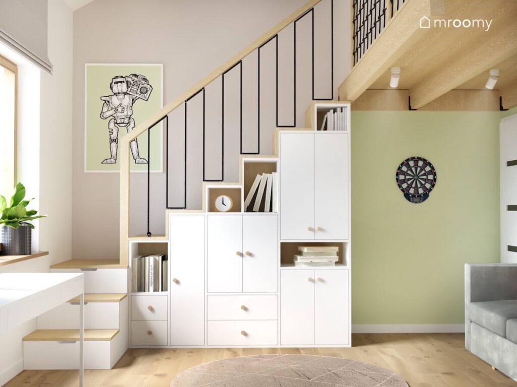 Biało drewniane meble wpasowane w schody prowadzące na antresolę a obok plakat z robotem i tarcza do darta w pokoju chłopca