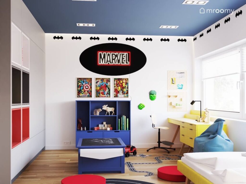 Biało niebieski pokój miłośnika Marvela i superbohaterów a w nim ciemnoniebieskie i żółte meble oraz liczne tematyczne dodatki