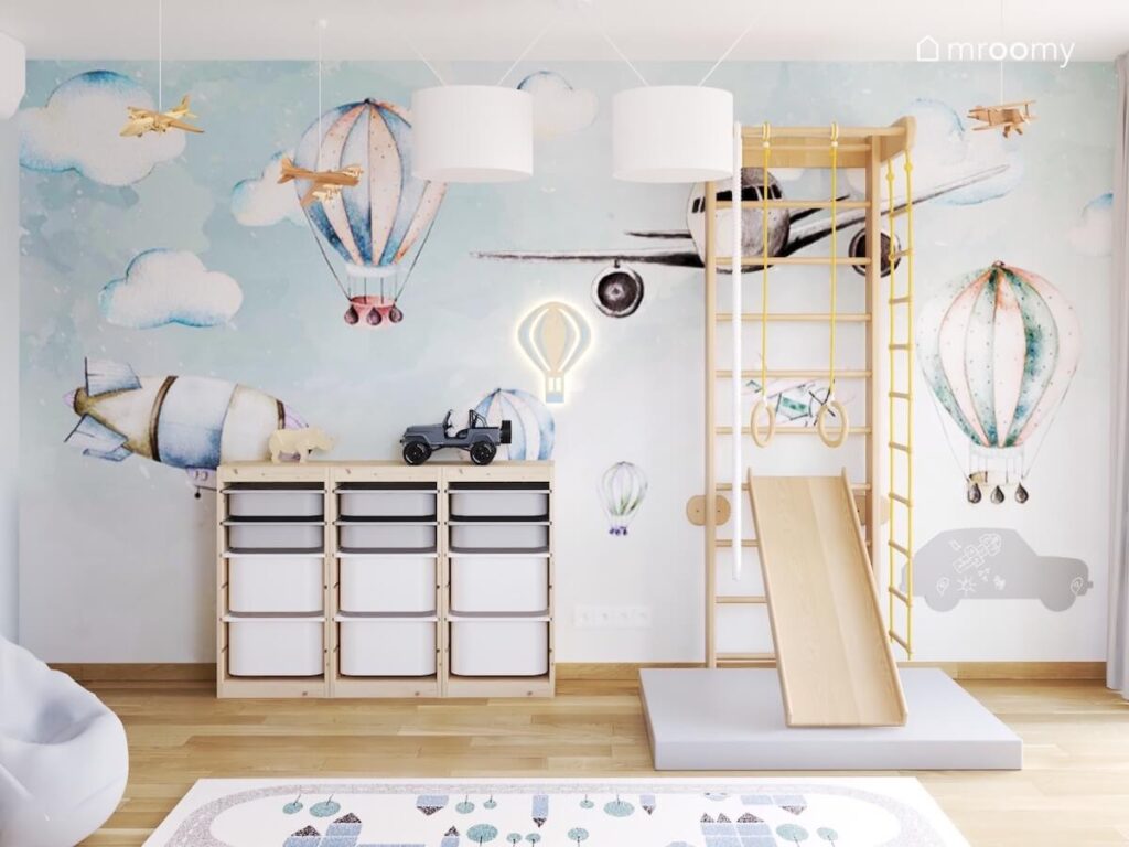 Ściana w pokoju rocznego chłopca pokryta tapetą w balony i samoloty a na niej drewniana drabinka gimnastyczna ze zjeżdżalnią i materacem oraz drewniany regał z pojemnikami