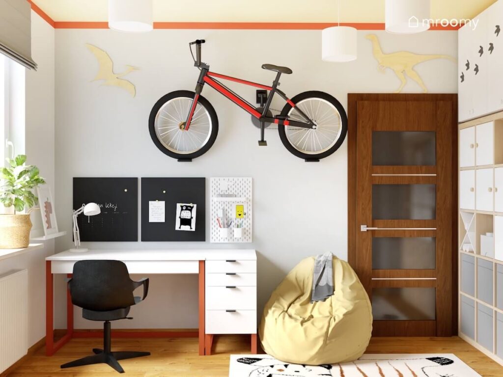 Strefa nauki w szarym pokoju dla chłopca obok jasnożółta pufa a na ścianie rower oraz sklejkowe dinozaury