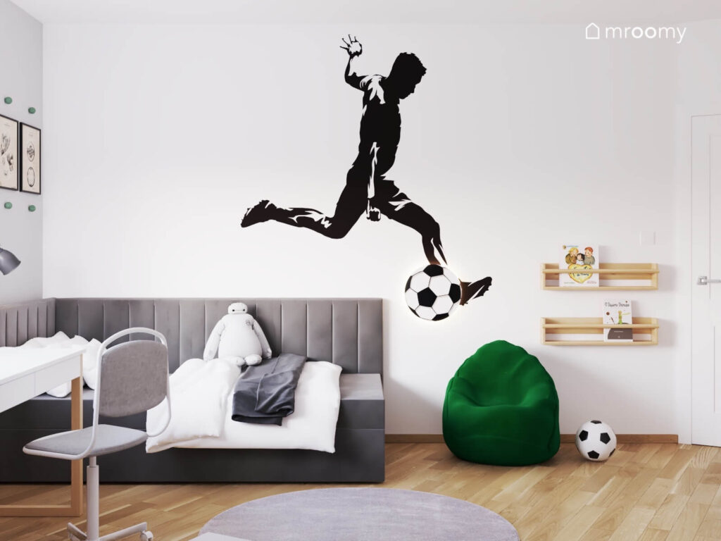 Biała ściana w pokoju chłopca z naklejką w kształcie piłkarza oraz lampką w kształcie piłki i drewnianymi półeczkami a pod nią szare łóżko i ciemnozielona pufa
