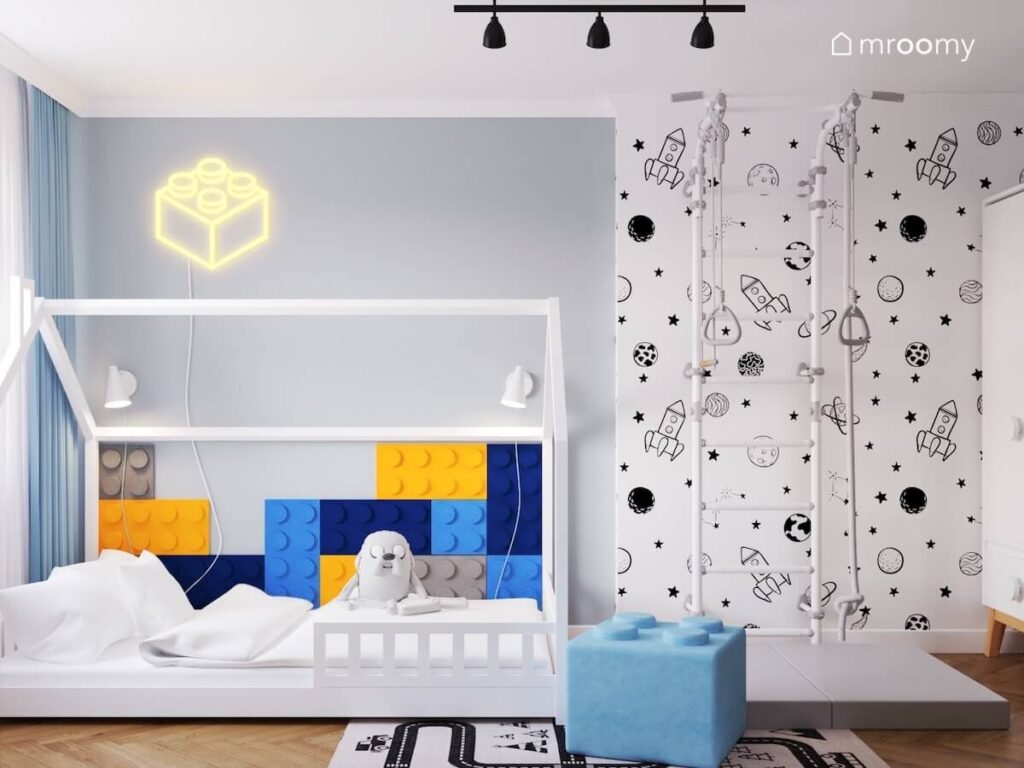 Białe łóżko domek uzupełnione kolorowymi panelami w kształcie klocków a nad nim ledon klocek a obok drabinka gimnastyczna i kosmiczna tapeta w pokoju chłopca