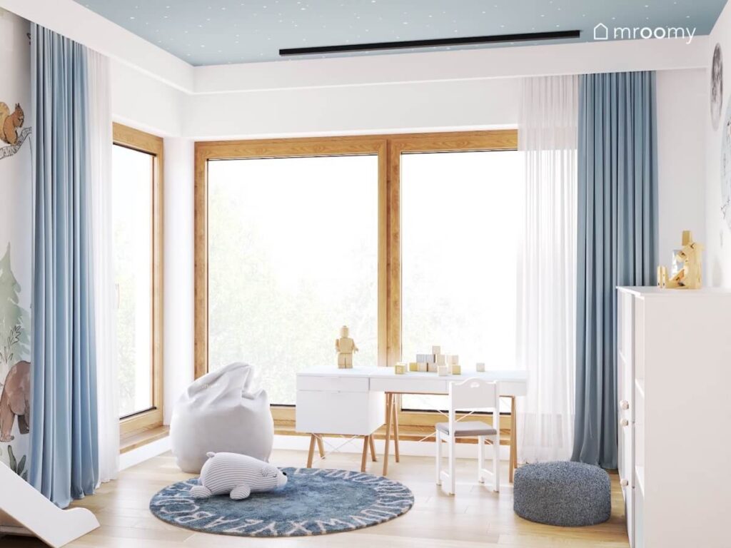 Biało niebieski pokój trzyletnich bliźniaków a w nim białe meble na drewnianych nogach i niebieskie dodatki w postaci dywanu zasłon i pufki
