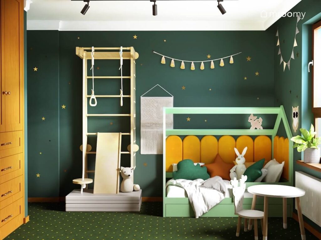 Ciemnozielona ściana w złote gwiazdki w pokoju dziewczynki a na niej drewniana drabinka gimnastyczna ozdobna makrama i girlanda pomponów a poniżej zielone łóżko domek uzupełnione żółtymi panelami ściennymi