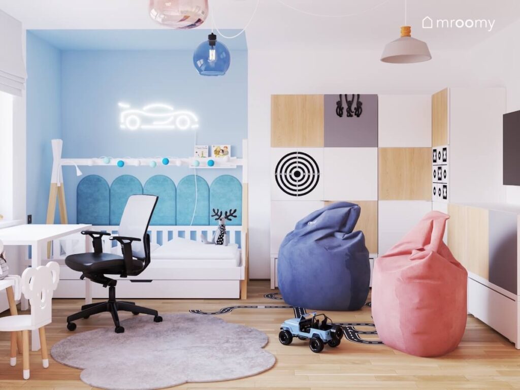 Niebieska strefa spania chłopca w pokoju dla rodzeństwa z białym łóżkiem uzupełnionym panelami ściennymi oraz girlandą cotton balls i ledonem w kształcie wyścigówki a obok szafa z nakładkami na fronty i dwie duże pufy
