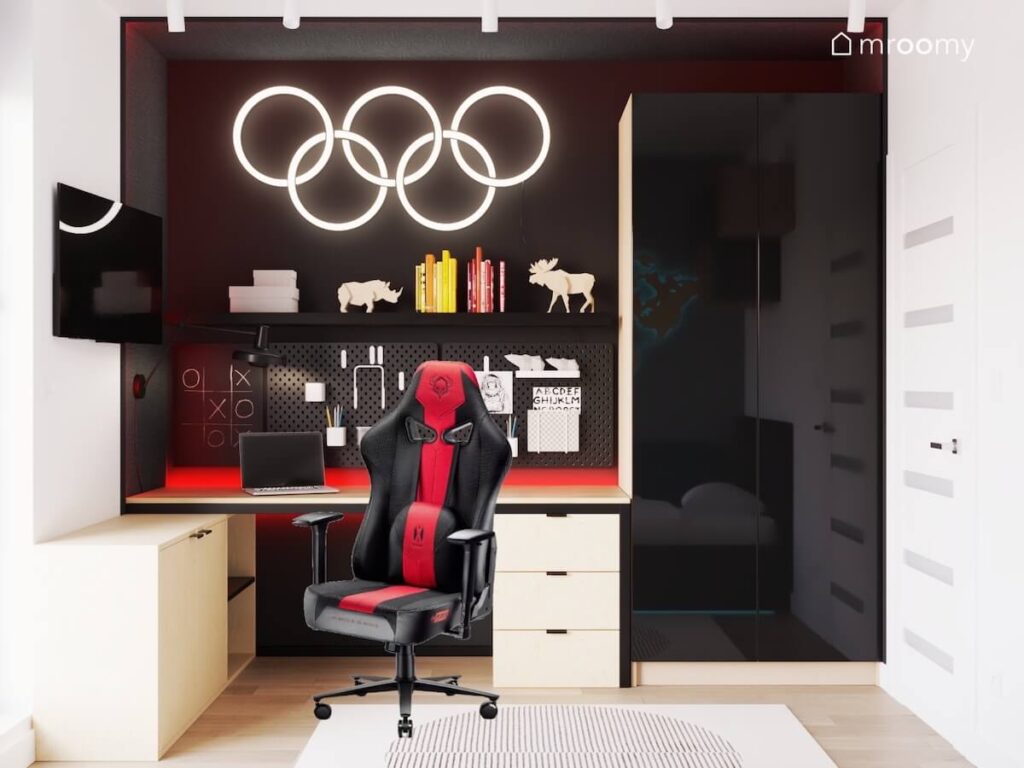 Ciemna strefa nauki w pokoju dziesięciolatka a w niej biurko kontener oraz fotel gamingowy a na ścianie półka organizery i ledon w kształcie symbolu olimpijskiego