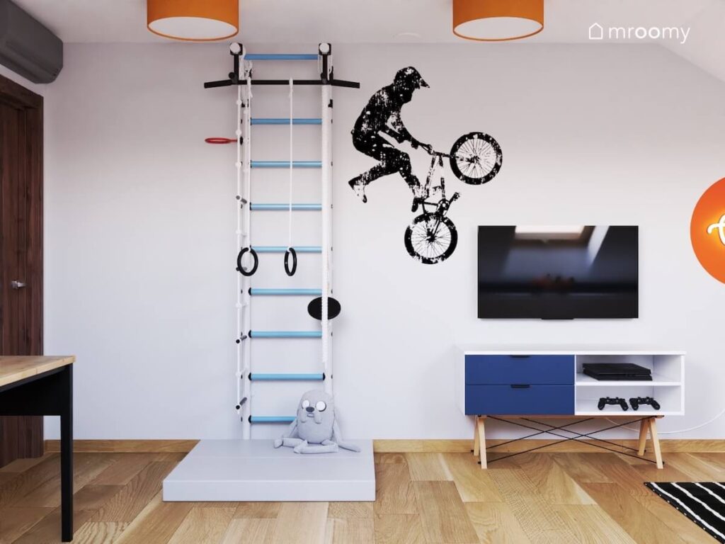 Biało niebieska drabinka gimnastyczna telewizor i szafka rtv a na ścianie naklejka z rowerzystą a na suficie pomarańczowe lampy w pokoju dla chłopca
