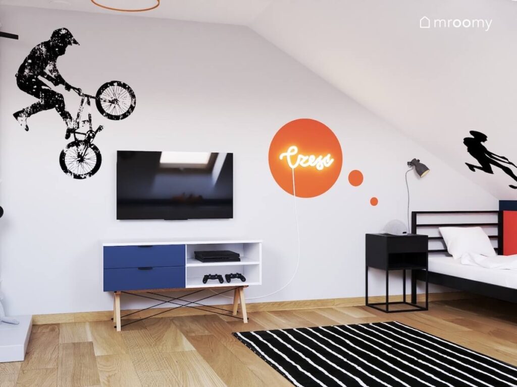 Biało szary poddaszowy pokój dla dziewięciolatka a w nim telewizor i szafka rtv ledon w kształcie napisu na pomarańczowym tle oraz czarne łóżko i szafka nocna