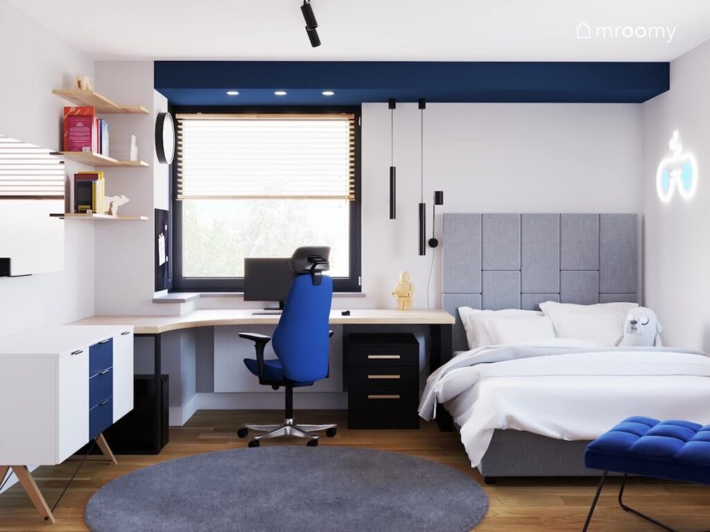 Jasny pokój dla dwunastolatka a w nim szare łóżko z wysokim zagłówkiem duże drewniane biurko na czarnych nogach oraz biało granatowa komoda