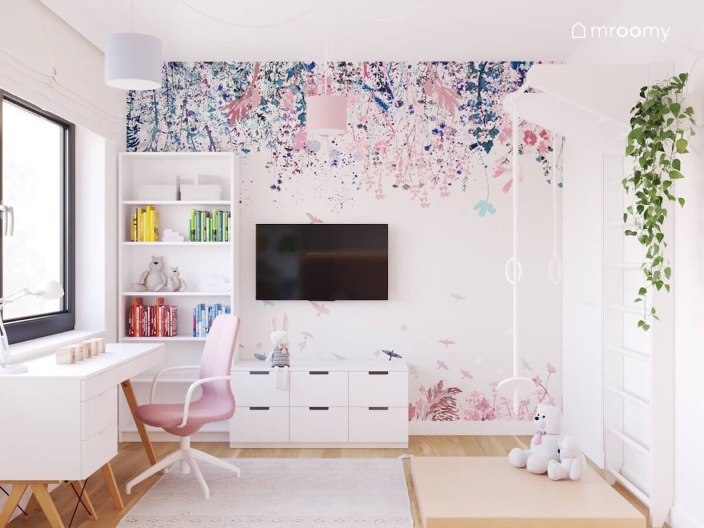 Ściana w pokoju siedmiolatki pokryta tapetą w różowo niebieskie kwiaty a na niej biały regał telewizor i niska szafka z szufladami