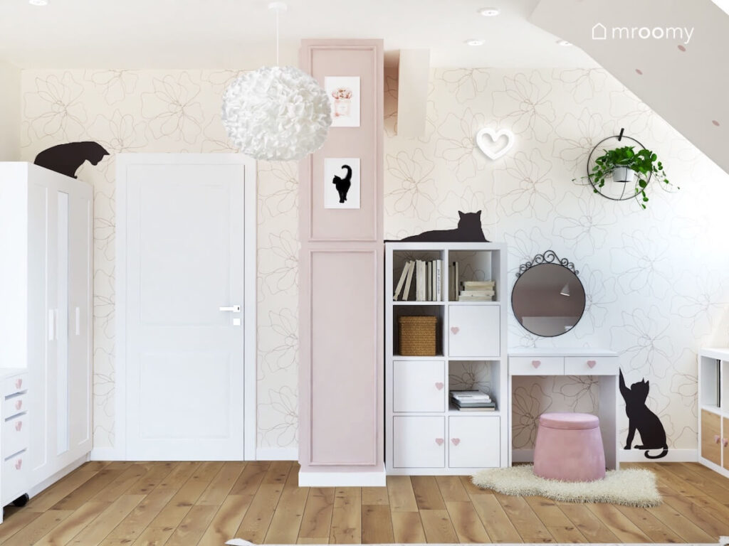 Ściana pokryta minimalistyczną tapetą w kwiaty w pokoju dziewczynki a na niej kwietnik ledon w kształcie serca i naklejki koty a pod nią toaletka z ozdobnym lustrem i biała szafka z gałkami w kształcie serc