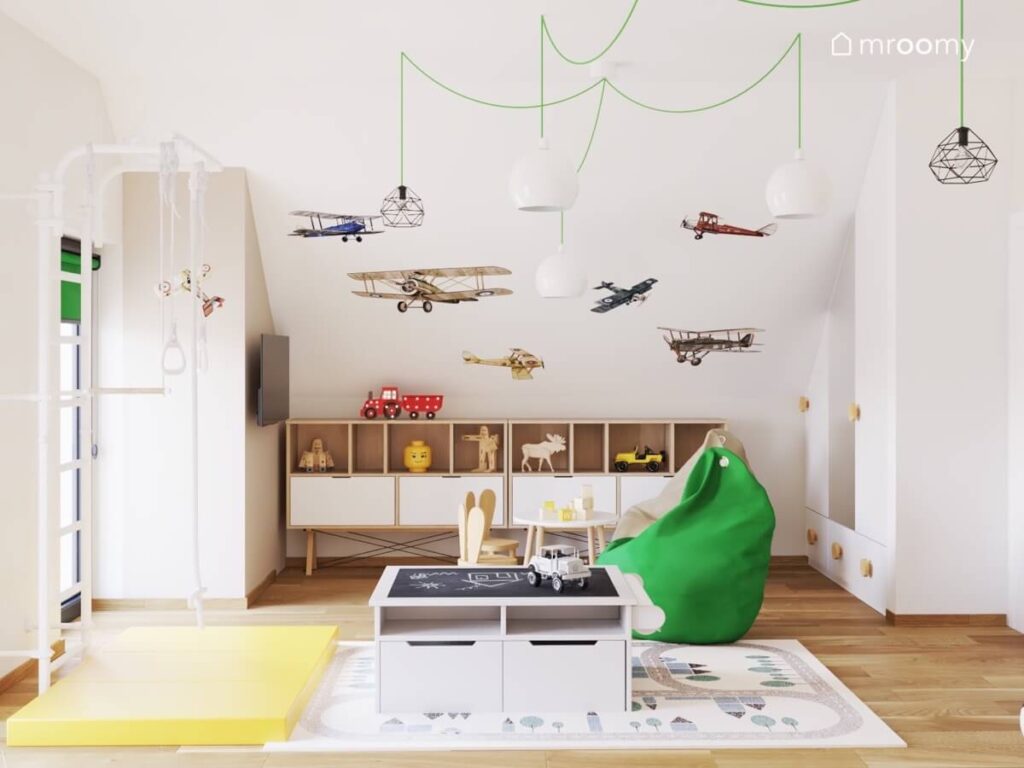 Jasny pokój dla trzylatka na poddaszu a w nim biało drewniane meble drabinka gimnastyczna zielona pufa oraz samoloty naklejki na skosie