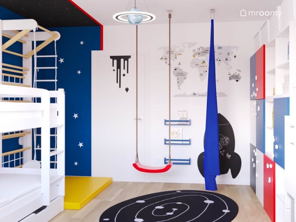 Biało niebieski pokój dwóch chłopców a w nim huśtawka wisząca szarfa mapa świata na ścianie tablica kredowa w kształcie rakiety oraz lampa w kształcie rakiety