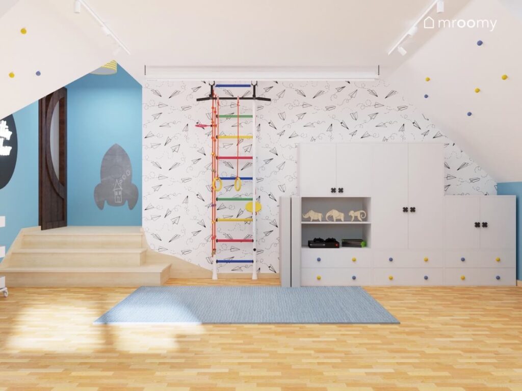 Ściana w pokoju dla chłopca pokryta tapetą w papierowe samoloty a na niej kolorowa drabinka gimnastyczna oraz białe meble modułowe z ozdobnymi gałkami