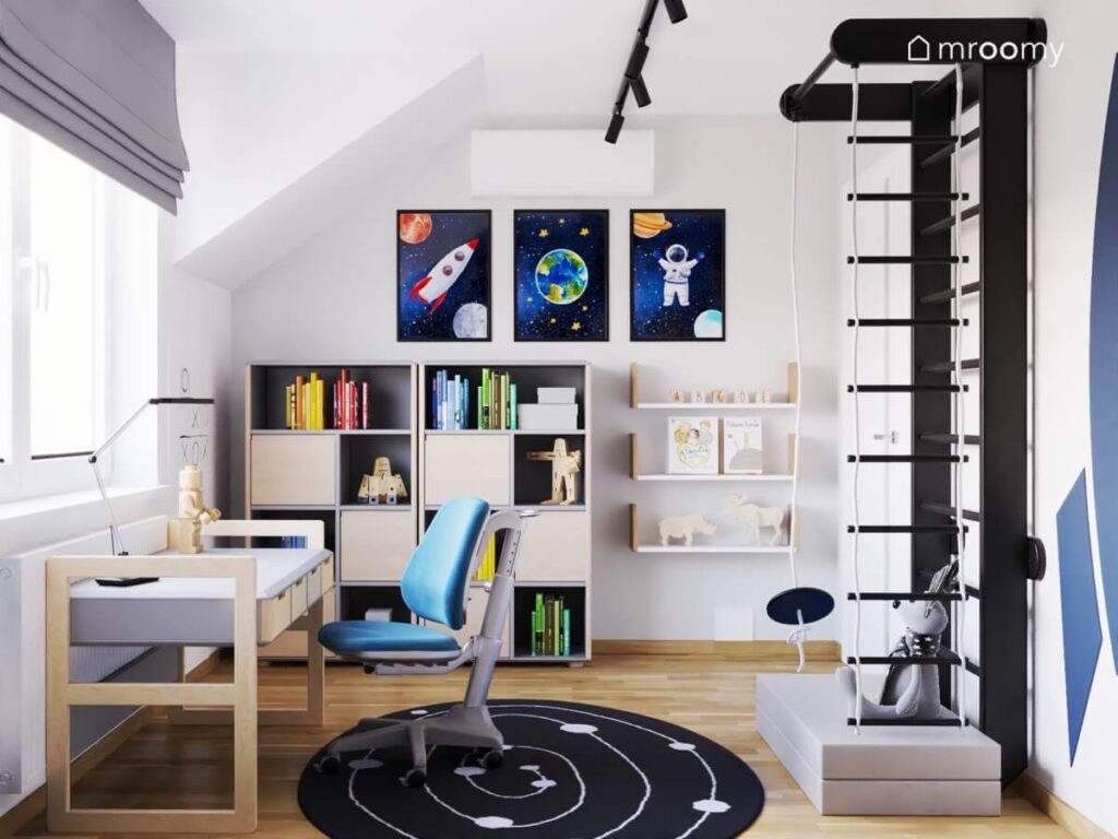Biały pokój dla chłopca na poddaszu a w nim szaro drewniane biurko regał z tej samej kolekcji kosmiczne obrazki na ścianie oraz czarna drabinka gimnastyczna