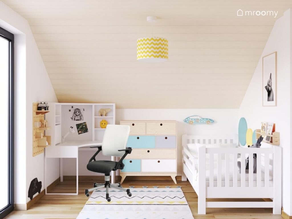 Biało beżowy poddaszowy pokój dla chłopca a w nim białe biurko kolorowa komoda oraz białe łóżko a na suficie biało żółta lampa