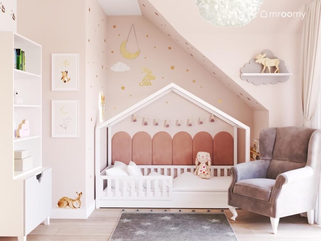 Białe łóżko domek uzupełnione panelami ściennymi oraz girlandą pomponów a nad nim złote gwiazdki księżyc i chmurka a obok półeczka chmurka i ozdobne plakaty w pokoju dziewczynki