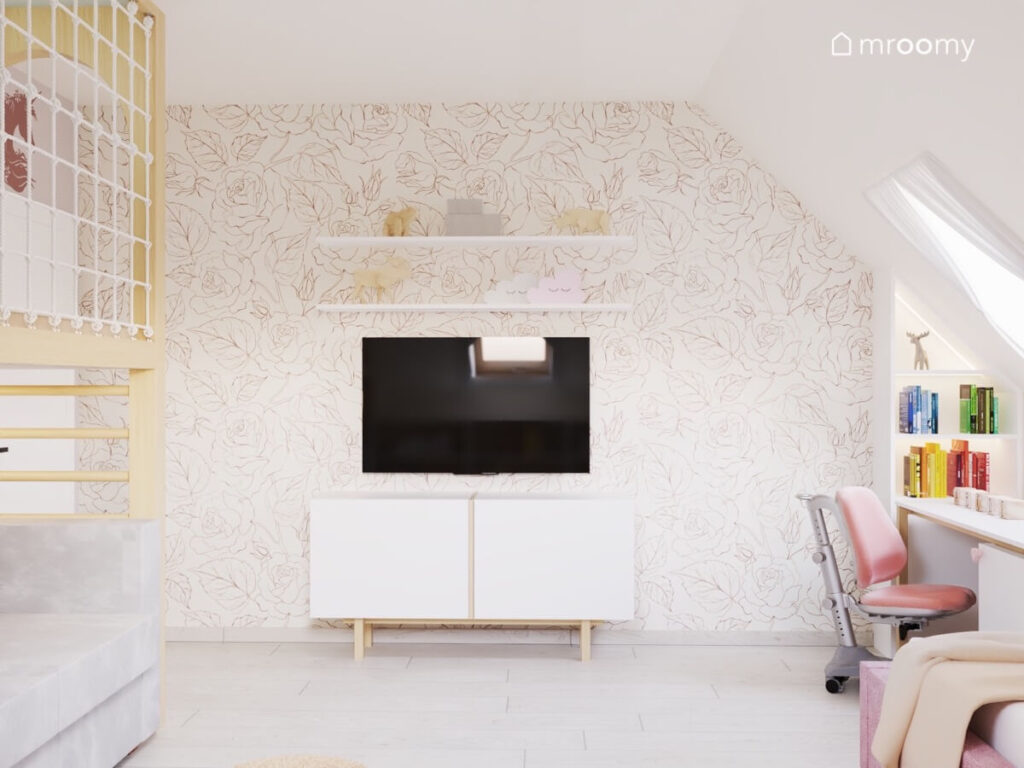 Ściana w poddaszowym pokoju dla dziewczynki a na niej tapeta w kwiaty telewizor półki oraz komoda