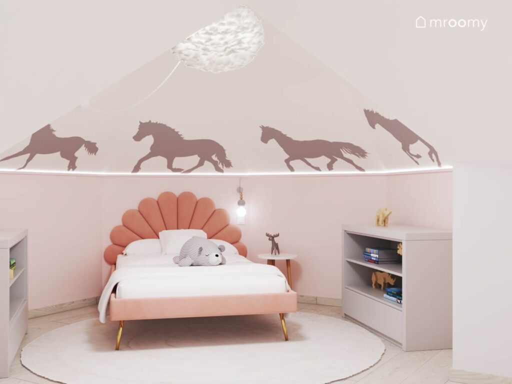 Strefa spania w pokoju dziewczynki w wieku szkolnym a w niej tapicerowane łóżko z ozdobnym wezgłowiem szafka na książki oraz naklejki ścienne w kształcie koni