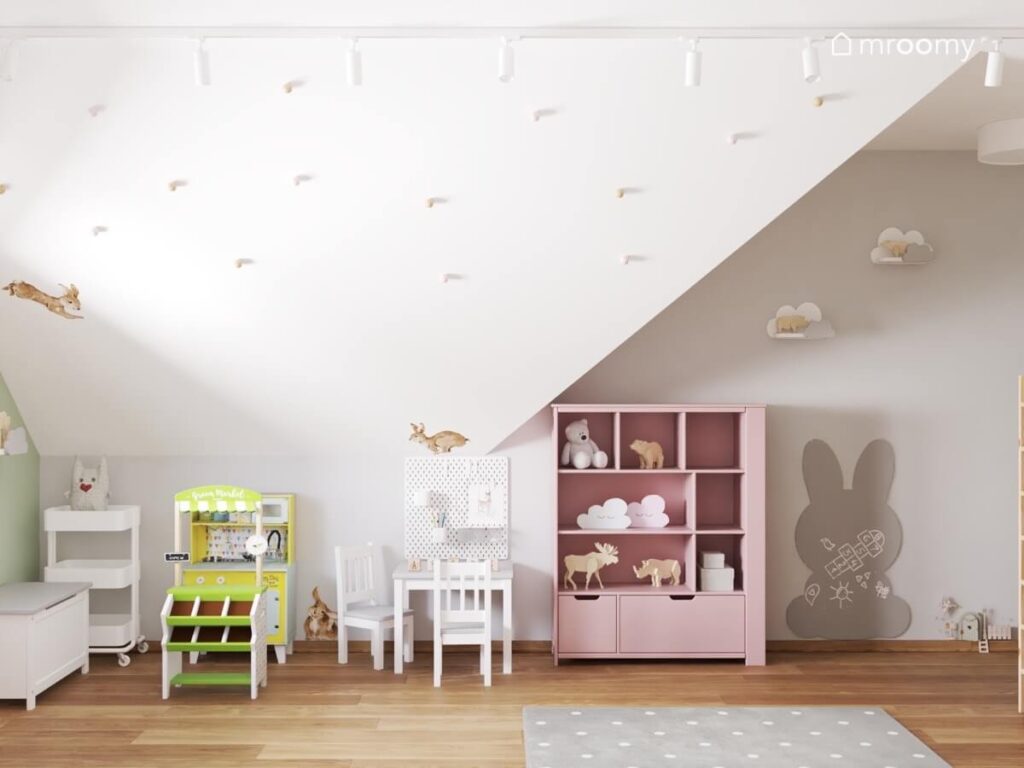 Ozdobiony kolorowymi gałkami i naklejkami w kształcie królików skos w pokoju małej dziewczynki a pod nim różowa szafka kuchnia dla dzieci i stolik z krzesełkami
