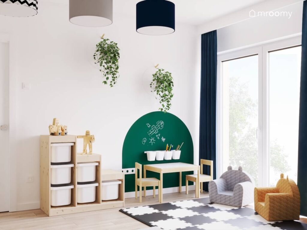 Drewniany regał z pojemnikami stolik z krzesełkami a na ścianie zielona powierzchnia kredowa i kwietniki w pokoju dwóch chłopców
