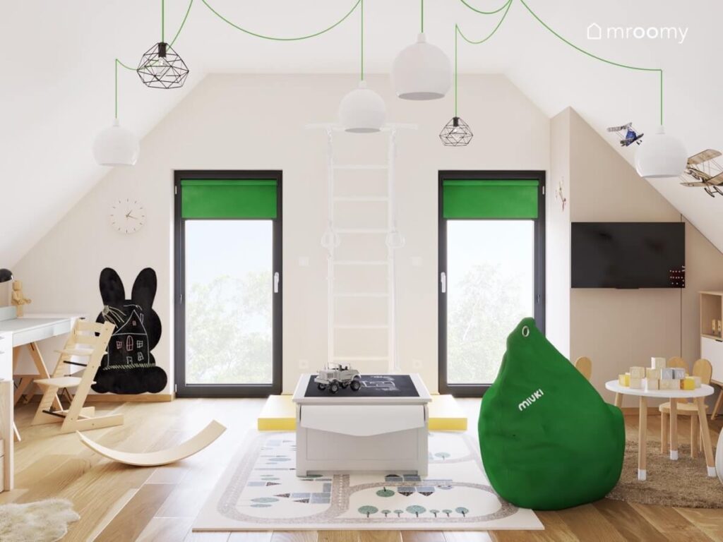 Przestronny poddaszowy pokój dla chłopca a w nim stolik do zabawy zielona pufa oraz rolety tablica kredowa w kształcie królika a na suficie lampy na rozłożystym zawieszeniu