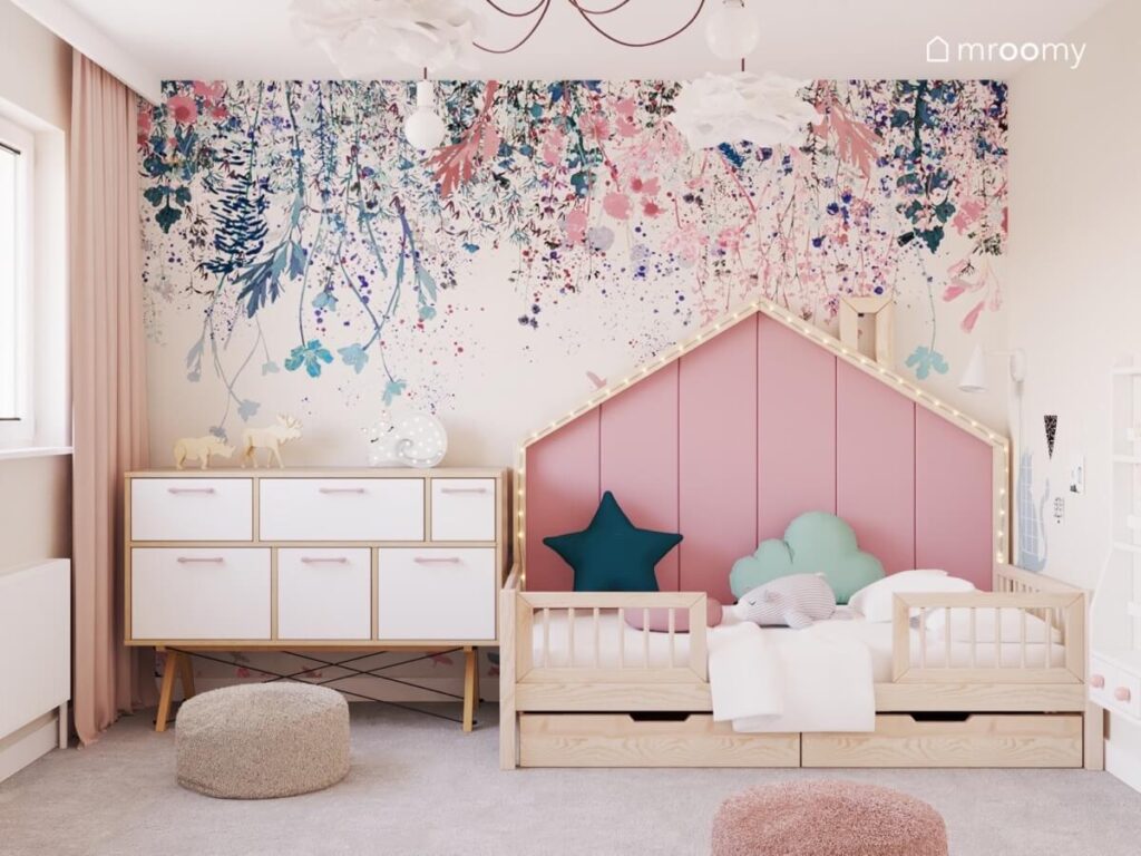 Drewniane łóżko domek w pokoju dla dziewczynki uzupełnione miękkimi panelami i światełkami obok biało drewniana komoda a na ścianie tapeta w kwiaty