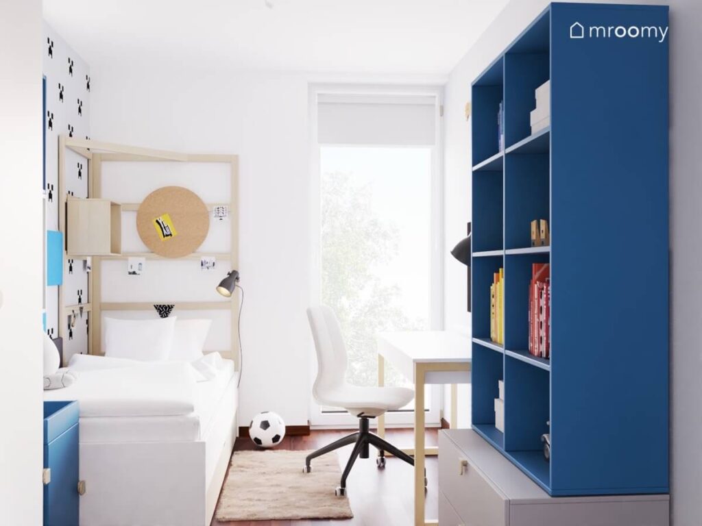 Jasny pokój dla chłopca w wieku szkolnym a w nim białe i drewniane meble oraz duży niebieski regał