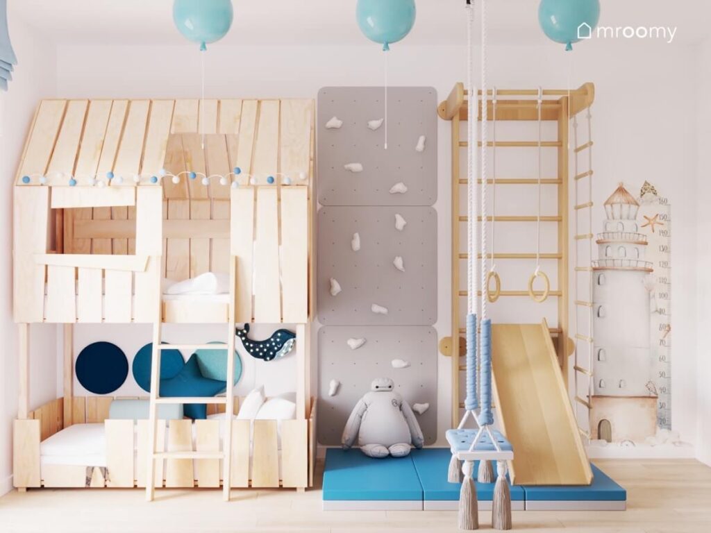 Drewniana antresola z miejscem do spania ścianka wspinaczkowa oraz drabinka gimnastyczna ze zjeżdżalnią i niebieskim materacem w pokoju rocznego chłopca