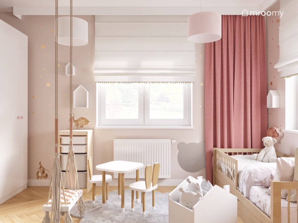 Beżowy pokój dla dziewczynki i chłopca a w nim biało drewniane meble huśtawka wisząca oraz różowe zasłony a na ścianie złote gwiazdki