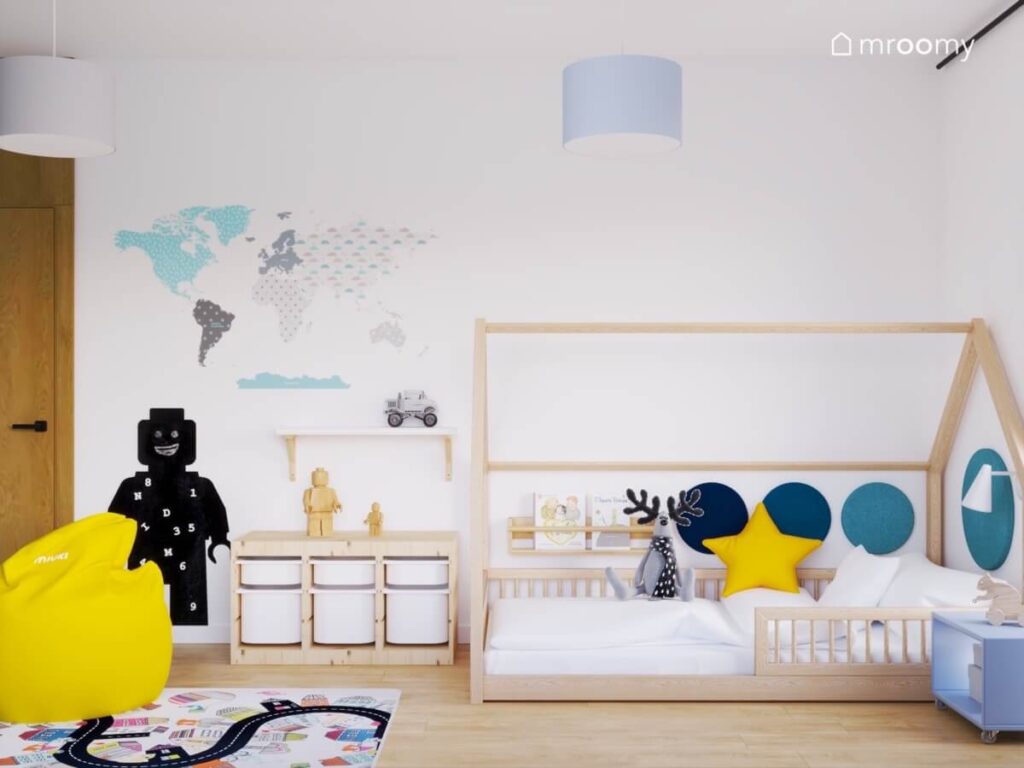 Drewniane łóżko domek uzupełnione niebieskimi panelami obok drewniany regał z pojemnikami mapa świata i tablica kredowa w kształcie ludzika Lego w pokoju małego chłopca
