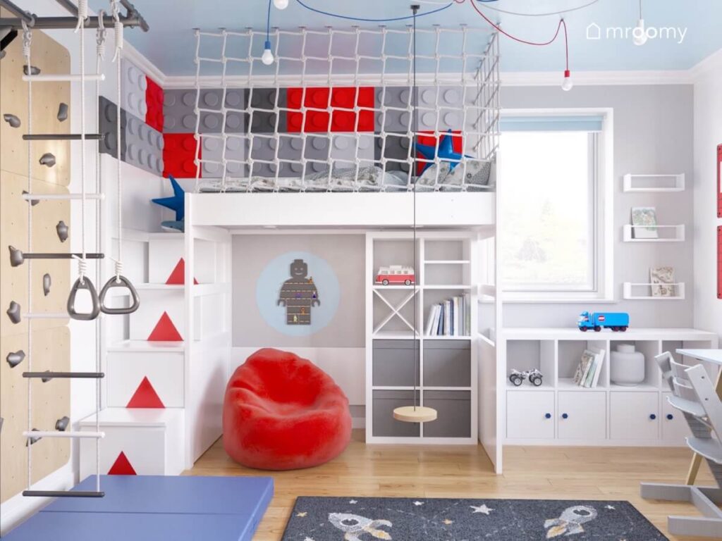 Wyłożona panelami w kształcie klocków antresola z miejscem do spania w pokoju kilkuletniego chłopca a pod nią regał półka w kształcie ludzika Lego i czerwona pufa